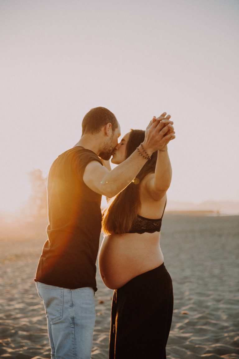 Babybauch- Paar Fotoshooting am Sandstrand. Paar steht sich gegenüber mit weit ausgebreiteten Armen, Sonne geht im Hintergrund unter
