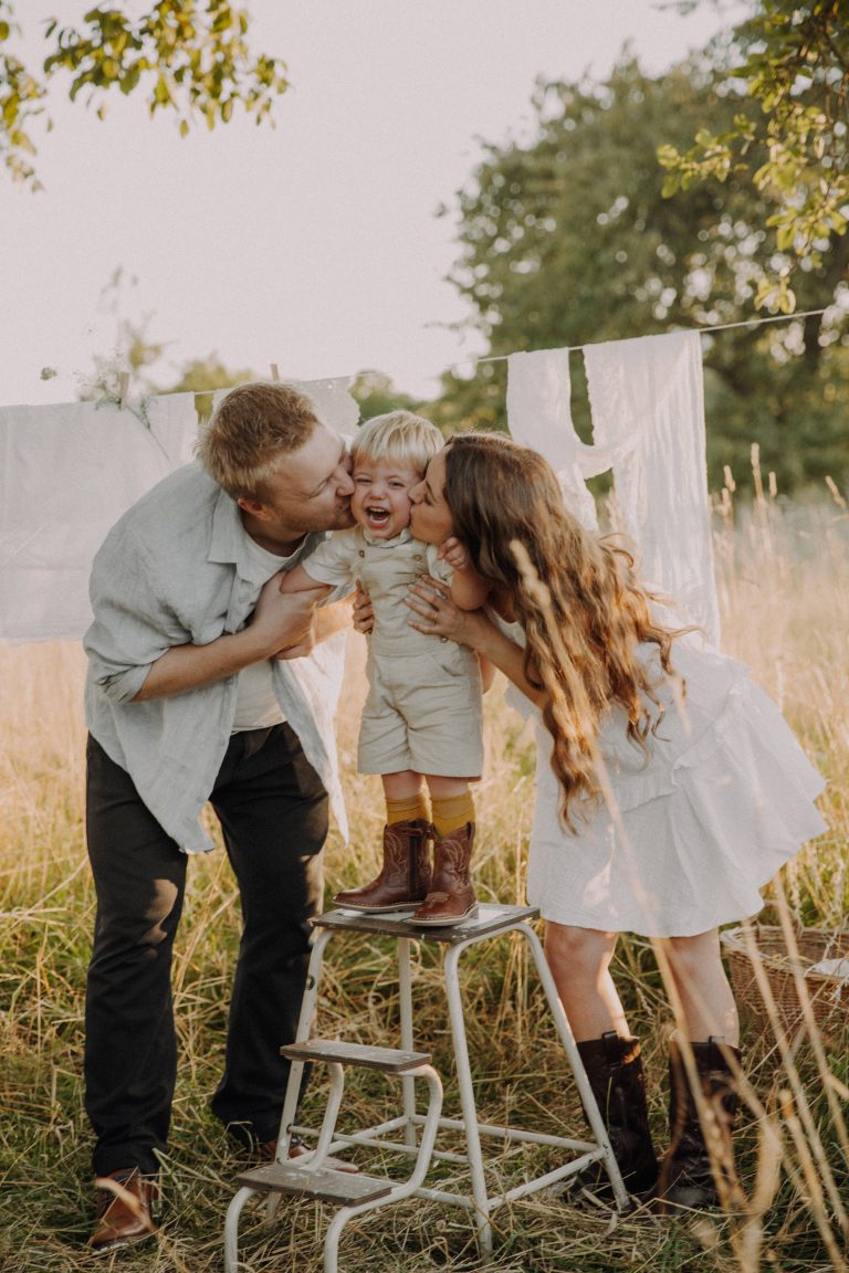 Familien Fotoshooting im Sommer – Eltern küssen ihr Kleinkind das auf Hocker steht, Wäscheleine und Sonnenuntergang im Hintergrund