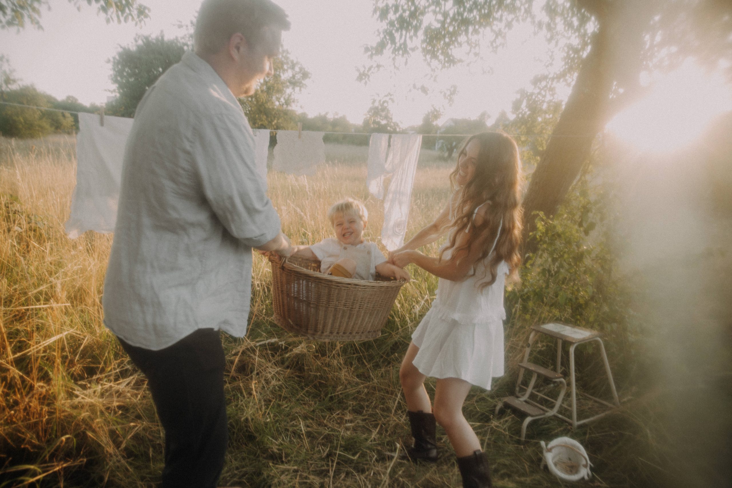 Mama und Papa schwingen ihren kleinen Sohn in einem Flechtkorb, alle lachen und haben Spaß auf einem Feld vor untergehender Sonne.