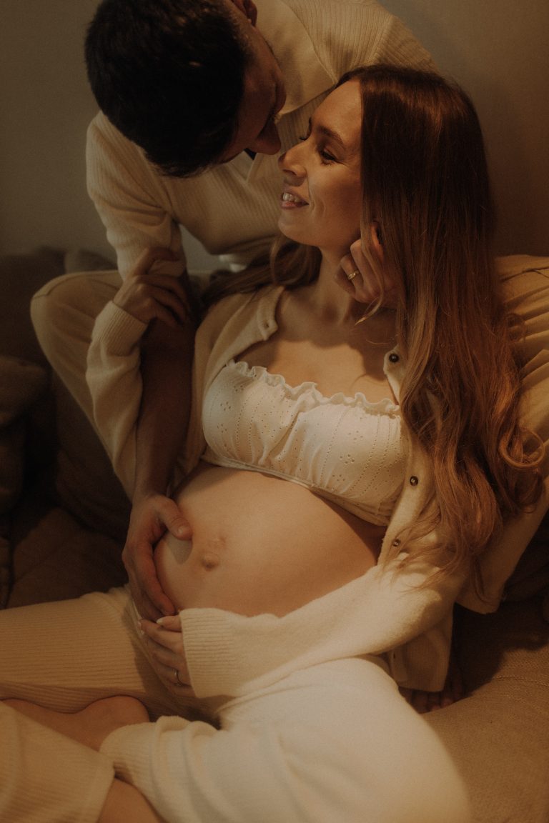 Emotionales Babybauch Paarshooting Indoor – schwangeres Paar in cremefarbenen Outfits in warmem Ambiente in ihrem Zuhause