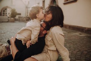 Familienfotoshooting in Altstadt. Familie sitzt auf dem Boden und kuschelt innig. Mama bekommt von Sohn einen Kuss. Die Sonne geht im Hintergrund unter