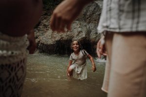Strahlendes Mädchen plantscht lachend im Wasser zwischen seinen Eltern – authentisches Familien Fotoshooting an einem Wasserfall in der Natur