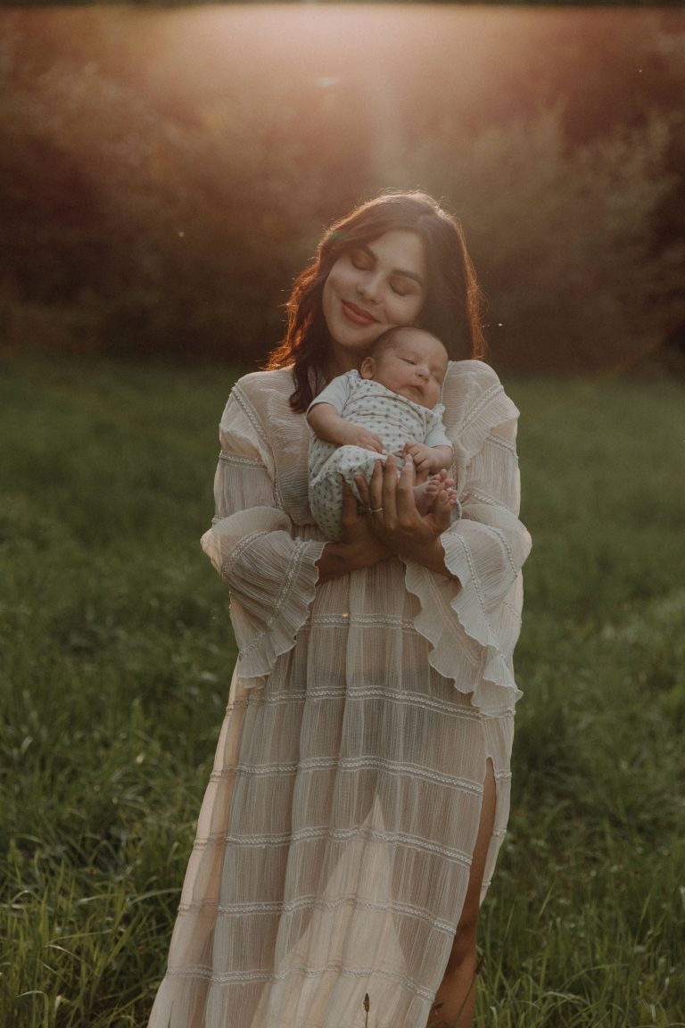 Mama in Maxikleid mit ihrem neugeborenen Baby auf dem Arm bei einem Fotoshooting auf grüner Wiese mit Sonnenuntergang im Hintergrund