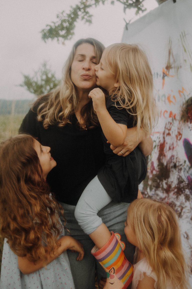 Mama-Kinder Fotoshooting im Freien. Mama spaziert mit ihren drei Töchtern über eine Wiese und kriegt einen Kuss auf die Wange.