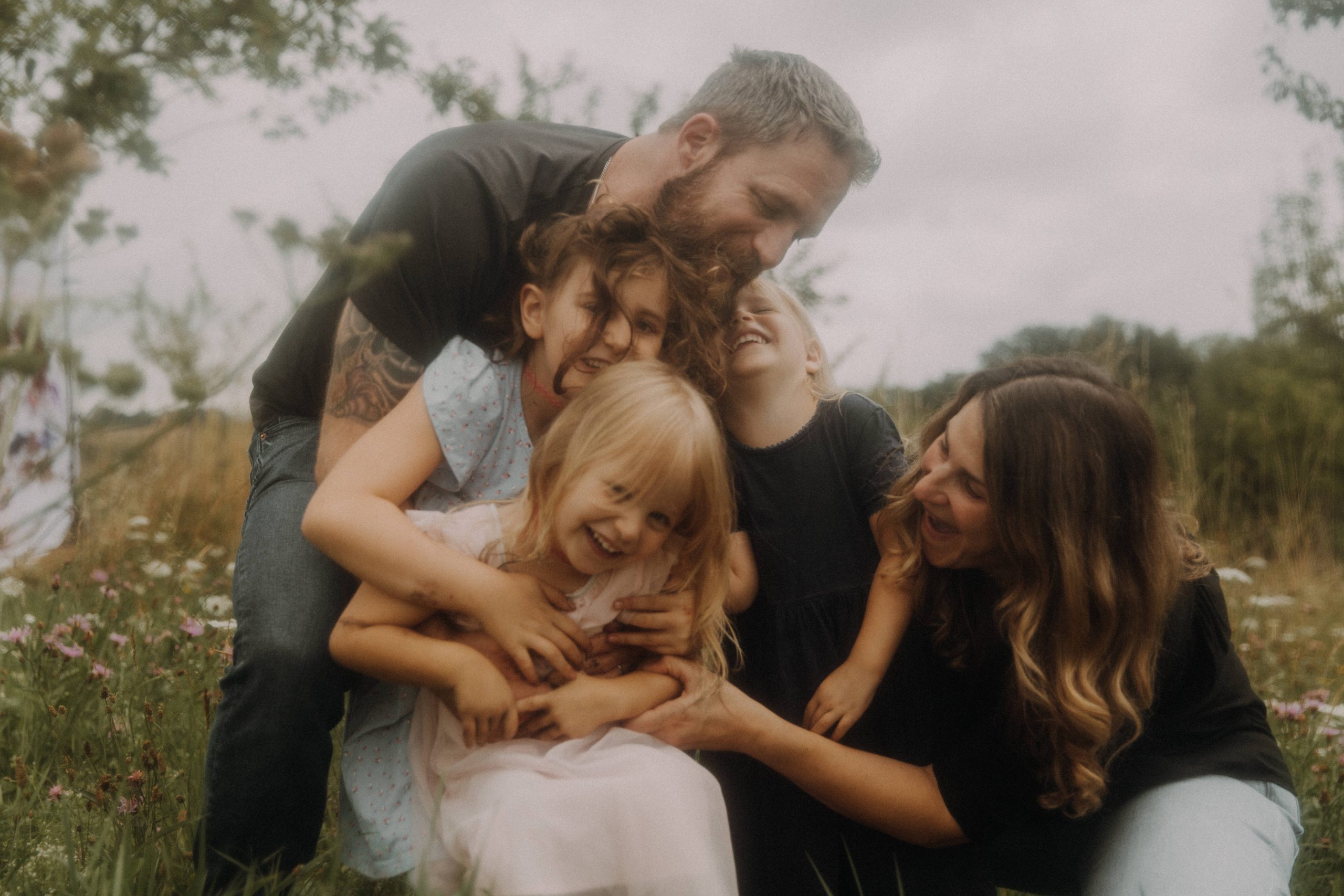 Lebendiges Familienfoto, Mama Papa und frei Töchter umarmen sich innig und lachend auf Blumenwiese
