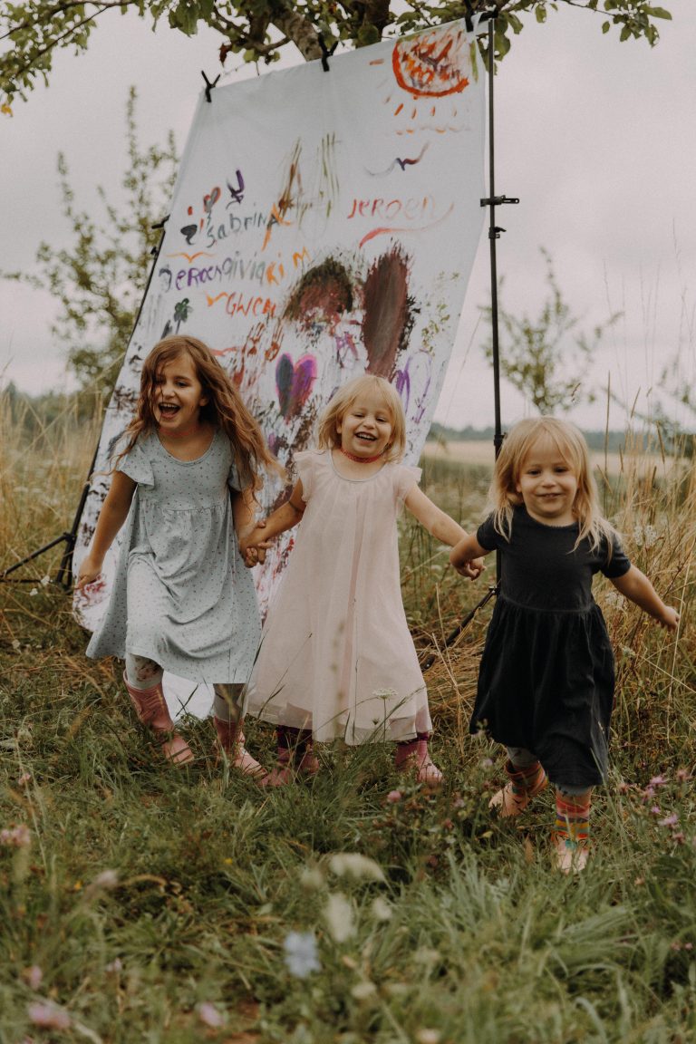 Erlebnisreiches Familien Fotoshooting Outdoor – drei lachende Mädchen mit selbst bemalter Leinwand auf der Wiese