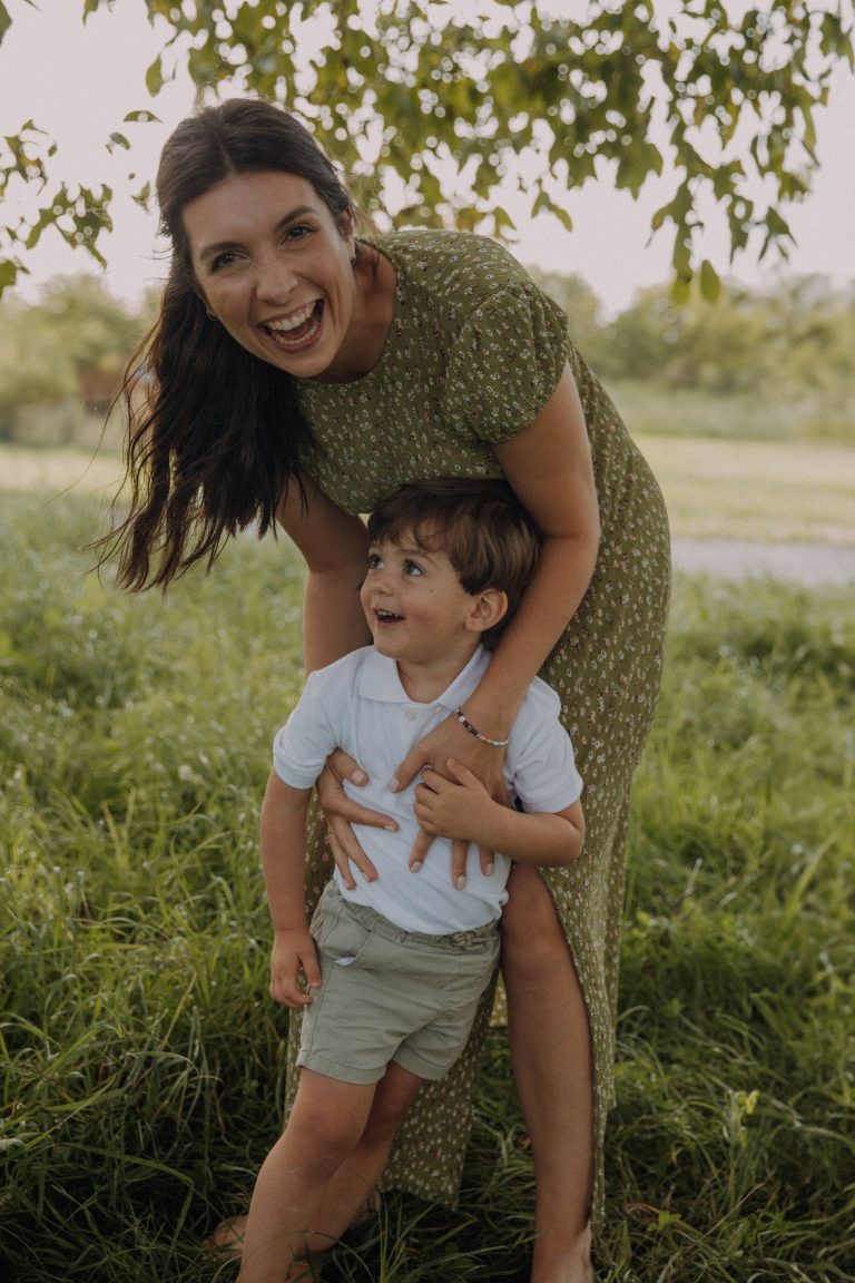 Mama-und Kind Fotoshooting auf einer grünen Wiese unter einem Nussbaum. Mama umarmt Sohn von hinten, beide lachen laut.