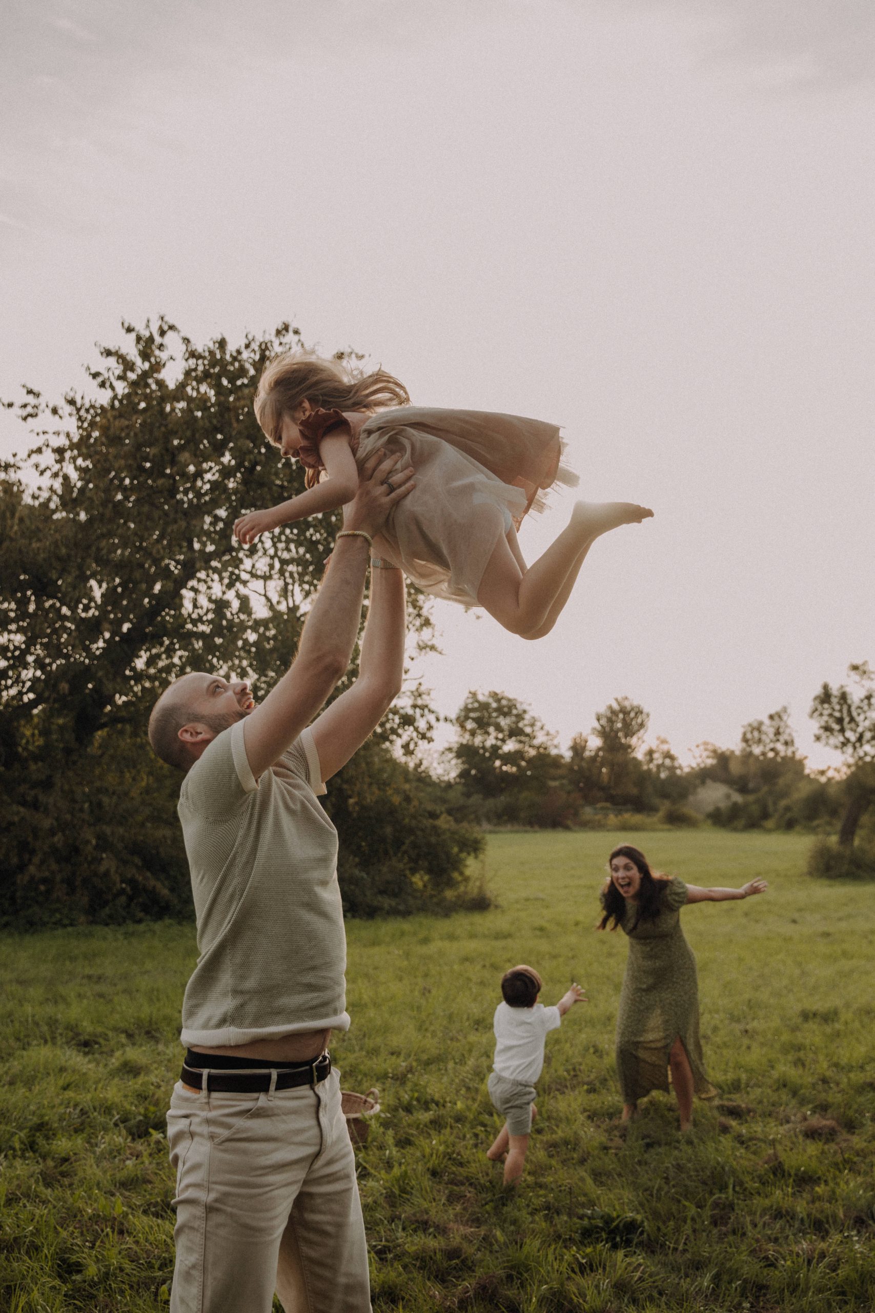 Authentisches Familien Shooting Sommer – Vater wirft Tochter in die Luft, im Hintergrund spielt die Mama mit dem Sohn auf grüner Wiese im Abendlicht