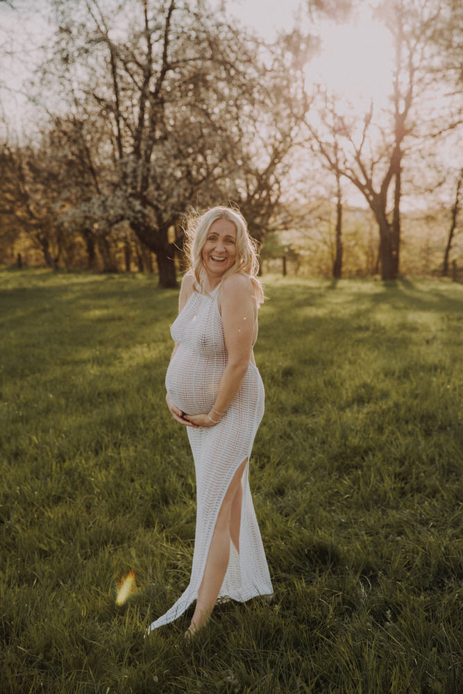 Outdoor Babybauch Shooting im Frühling – schwangere Frau in enganliegendem weißem Strickkleid auf Wiese im goldenen Abendlicht