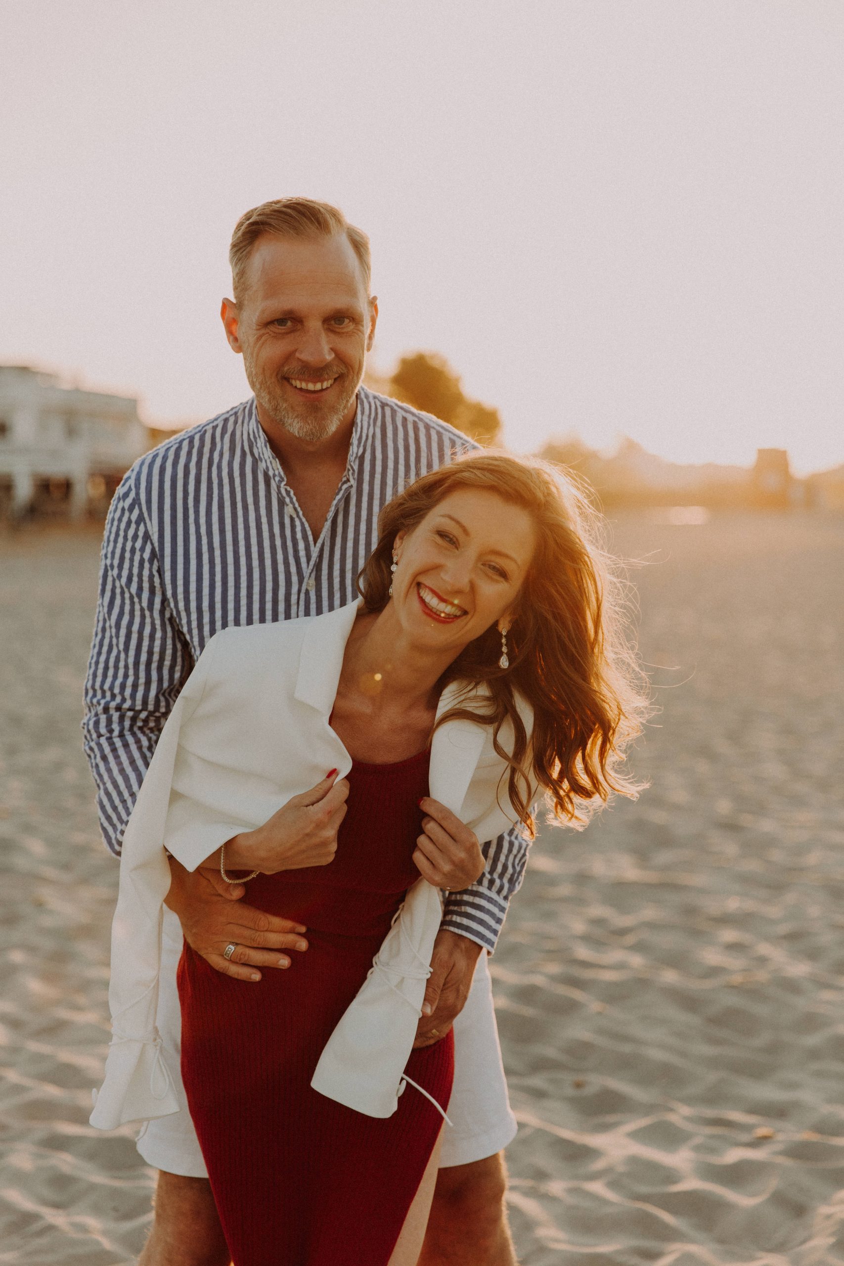 Glückliches Paar umarmt sich lachend am Strand im Sonnenuntergang bei einem Paarshooting
