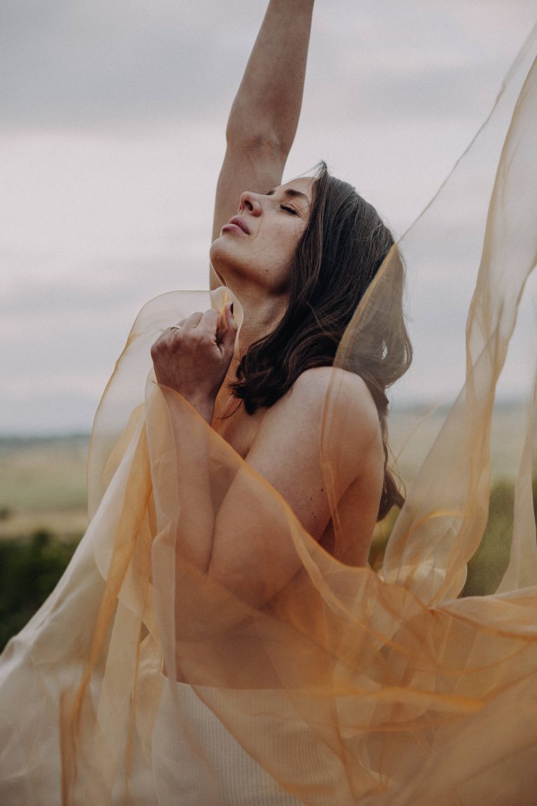 Fine Art Boudoir Shooting – Frau mit fließendem goldenen Tüll im Wind, dramatischer Himmel im Hintergrund