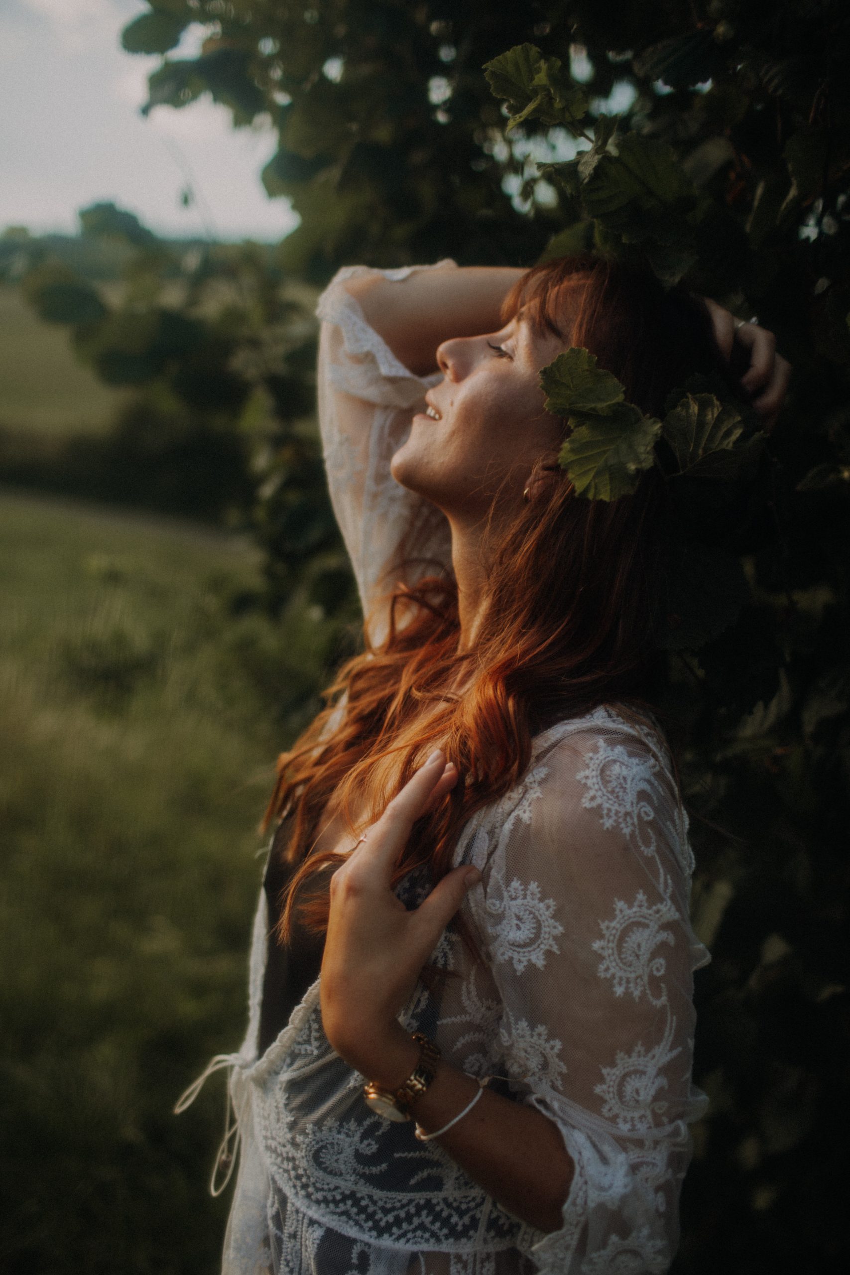 Frau mit langen roten Haaren im weißen Boho-Spitzenkleid lehnt an einem Baum und blickt träumerisch in den Himmel – sinnliches Boudoirshooting im Abendlicht