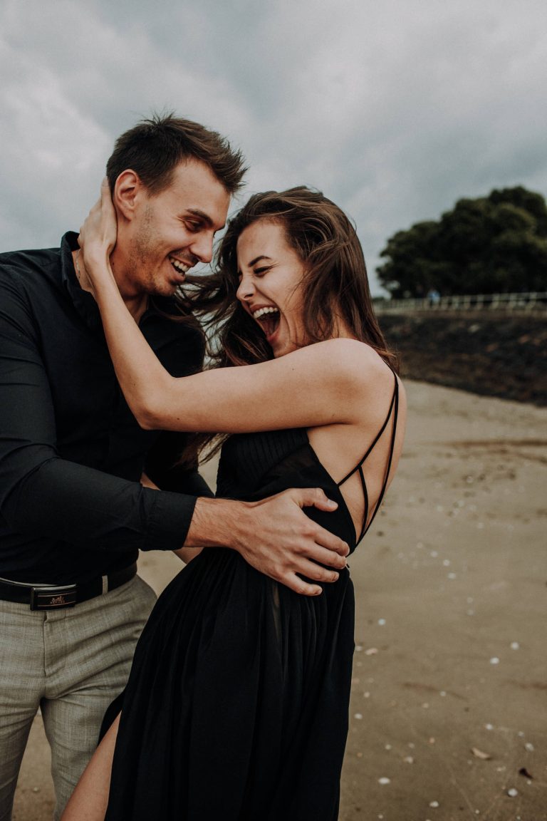 Frau im schwarzen Rückenfreien Kleid und Mann im dunklen Hemd lachend beim Paarshooting am Sandstrand