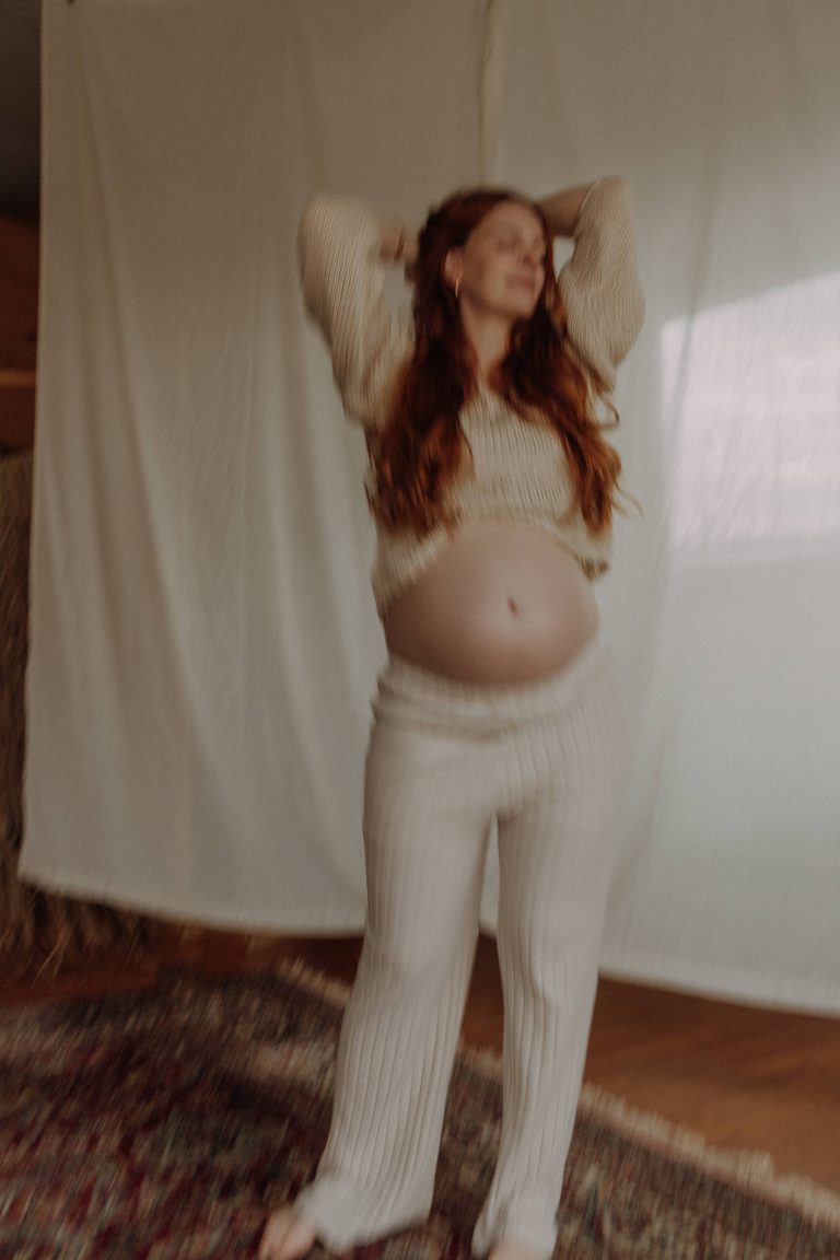 Babybauch Shooting im Film Look Indoor – schwangere Frau in cremefarbenem Strick-Outfit im stimmungsvollen natürlichen Licht
