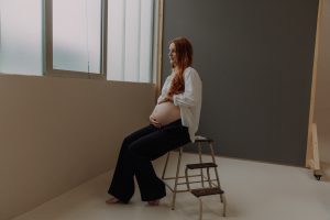 Schwangere Frau mit roten Haaren sitzt barfuß auf einem Hocker und hält ihren Babybauch – minimalistisches Schwangerschafts Fotoshooting im Studio