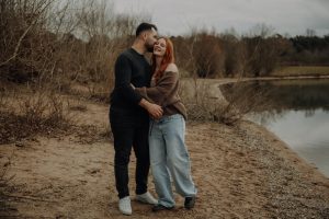 Verliebtes Paar umarmt sich lachend an einem Seeufer im Herbst – authentisches Babybauch Fotoshooting in der Natur