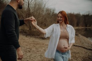 Schwangere Frau mit roten Haaren lacht strahlend, während ihr Partner sie an einem Seeufer dreht – lebendiges Babybauch Paar Fotoshooting in der Natur