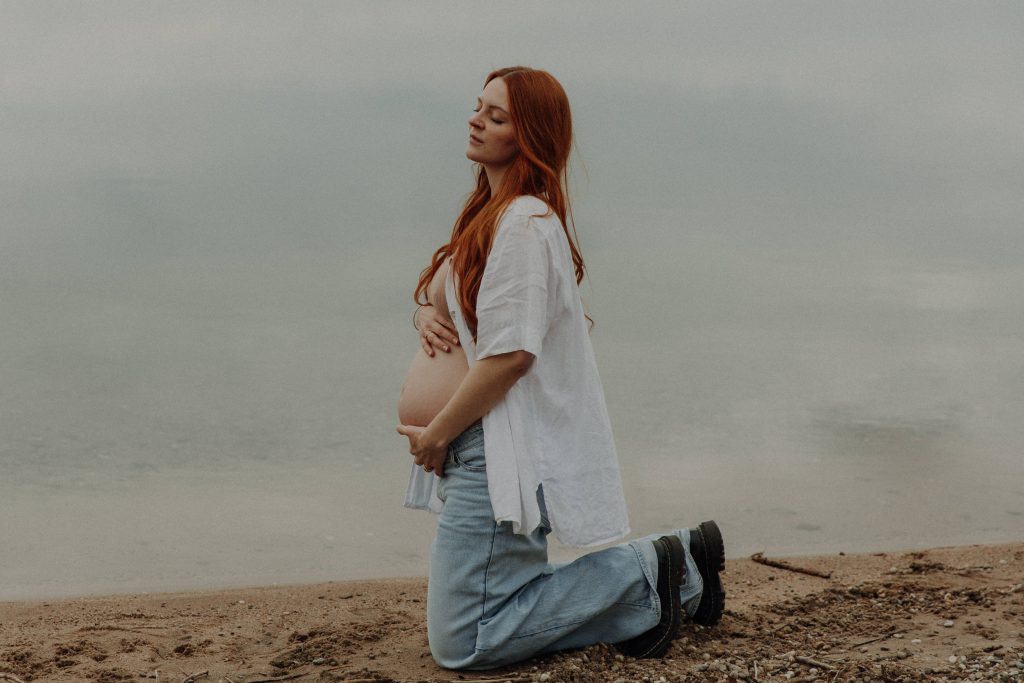 Sinnlich, kunstvolles Foot einer Schwangeren vor dem See. Sie kniet im Hand und hält ihren Babybauch