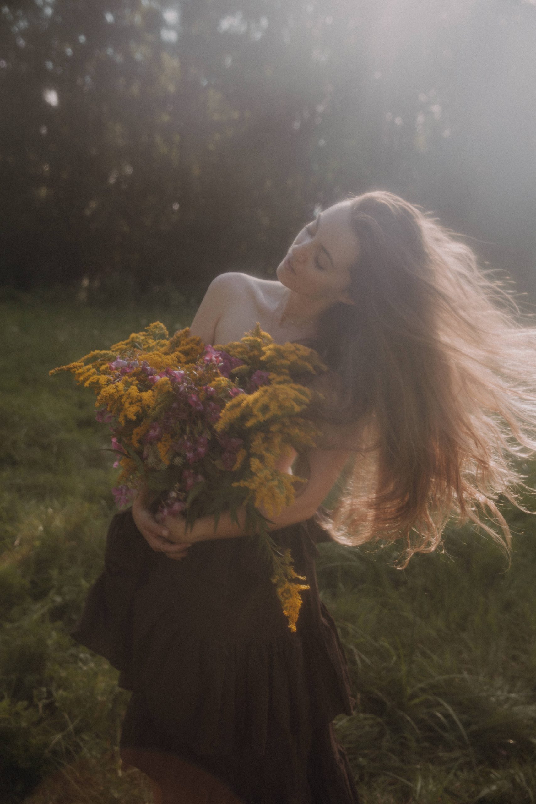 Märchenhaftes Outdoor Boudoir Shooting – Frau mit wehenden Haaren hält Wildblumenstrauß im sanften Gegenlicht der Natur