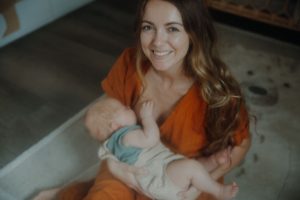 Strahlende Mutter mit langen braunen Haaren hält ihr Neugeborenes im Arm – authentisches Newborn Fotoshooting zu Hause