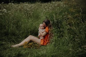 Mutter sitzt barfuß im hohen Wiesengrass und kuschelt ihr Baby – natürliches Newborn Familienshooting im Freien