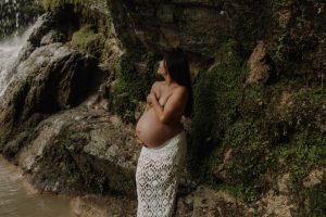Schwangere Frau lehnt an einer moosbedeckten Felswand neben einem Wasserfall und hält ihren Babybauch – stimmungsvolles und natürliches Babybauch Fotoshooting in der Natur