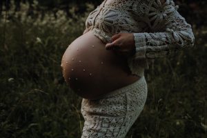 Nahaufnahme eines Babybauchs mit kleinen Perlen verziert, schwangere Frau im Häkelkleid auf einer Wiese – kreatives und stimmungsvolles Babybauch Fotoshooting in der Natur