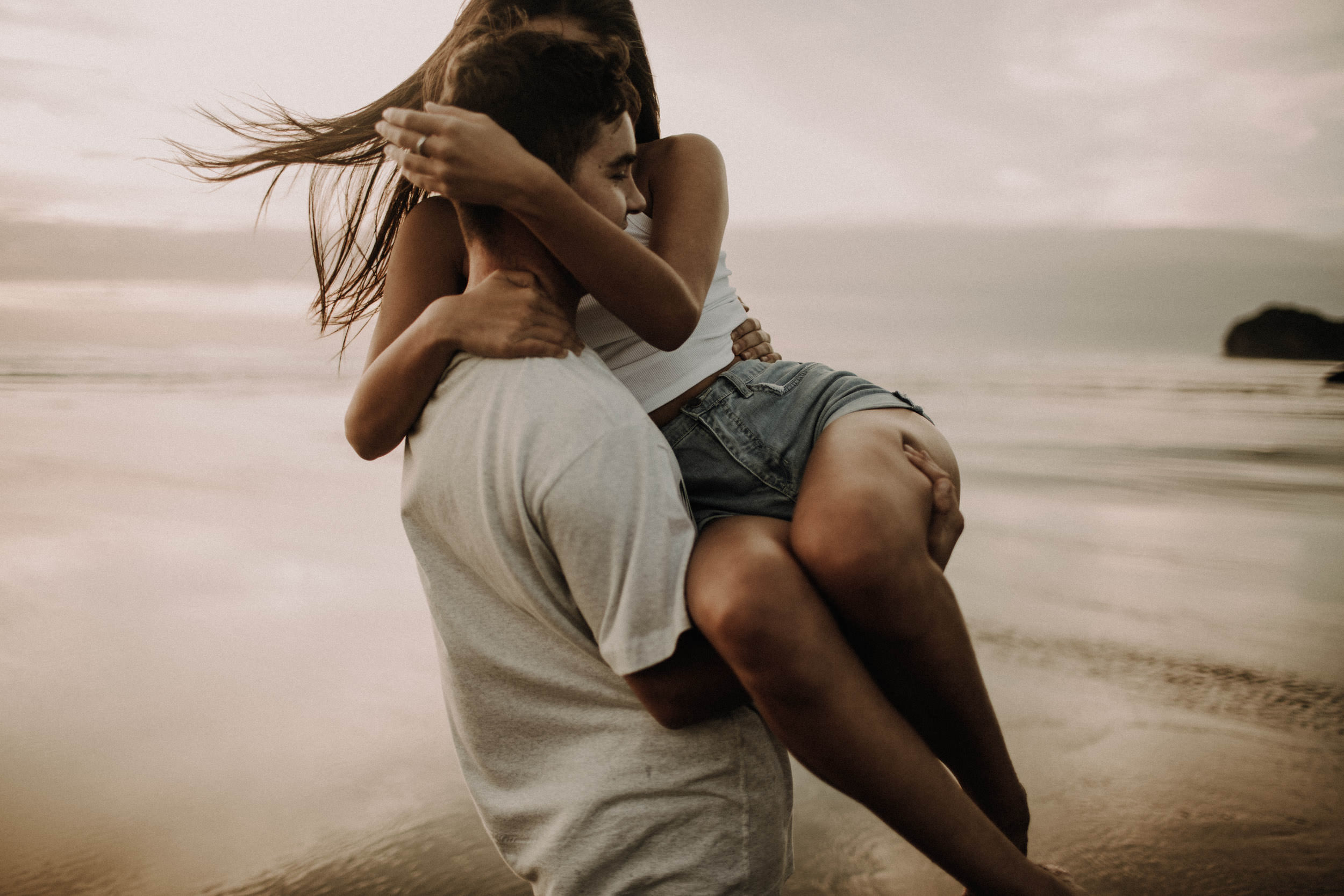 Leidenschaftliches Couple Shooting am Strand – Mann hebt Partnerin hoch in innigem Kuss-Moment am Wasser