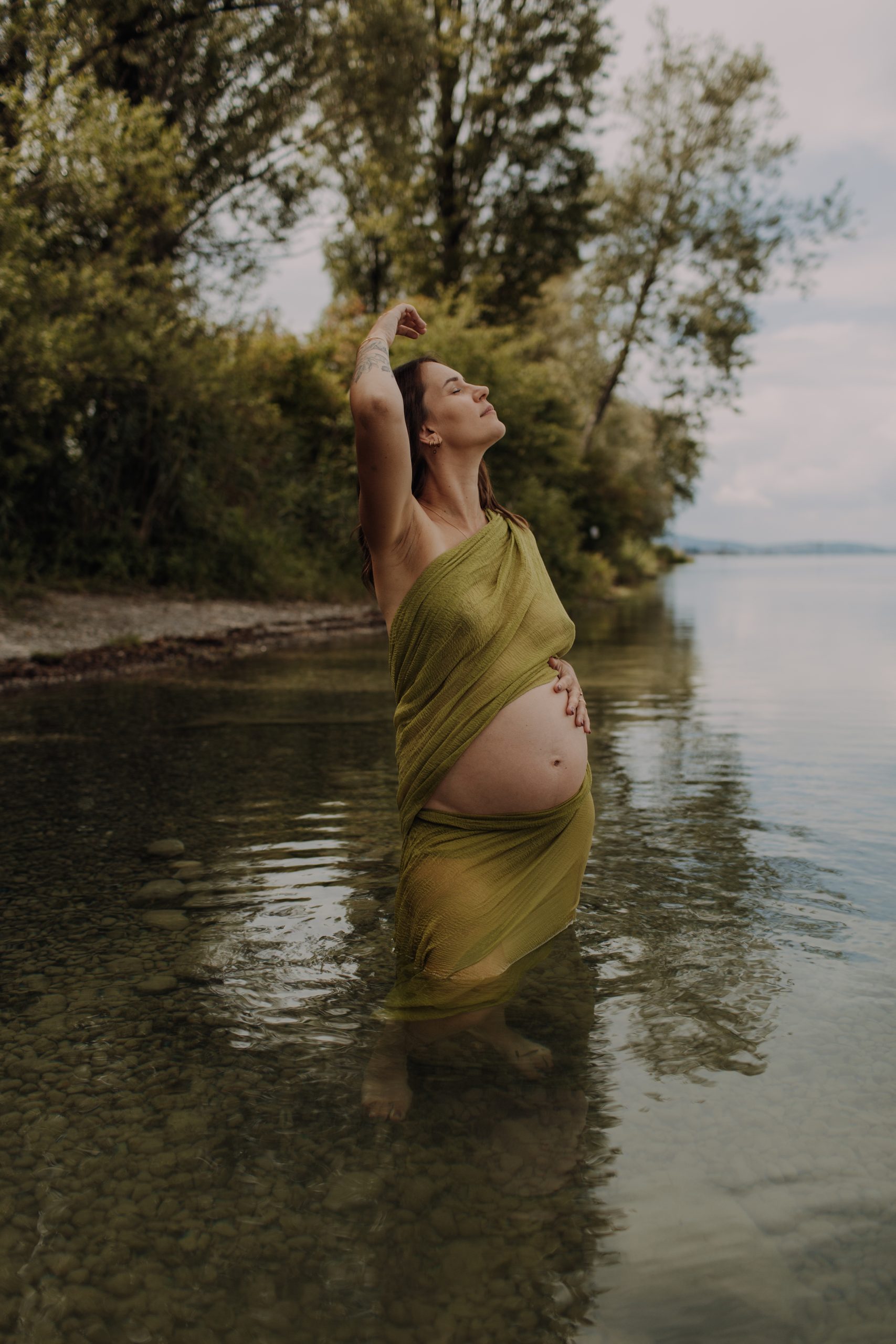 Outdoor Babybauch Shooting am Bodensee – schwangere Frau in olivgrünem Tuch im Wasser, umgeben von Natur