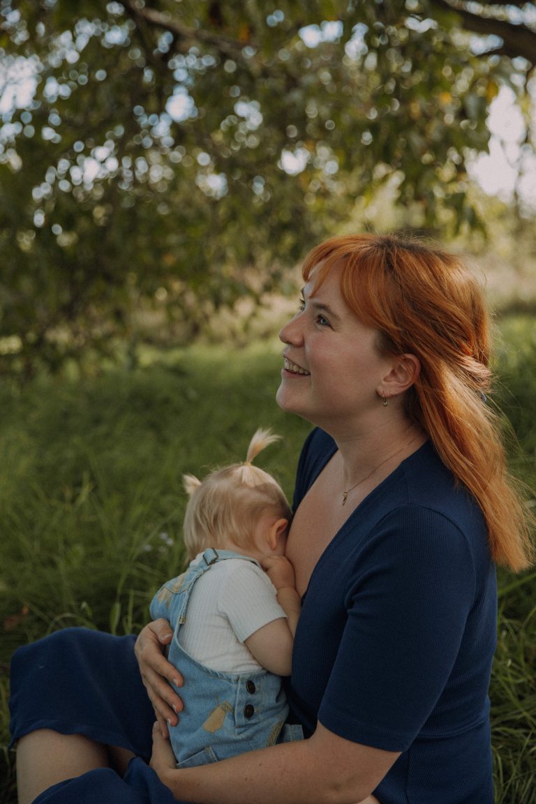 Rothaarige Mama stillt ihre Tochter unter einem Nussbaum im sommerlichen Abendlicht bei einem Mutter- und Kind Fotoshooting.