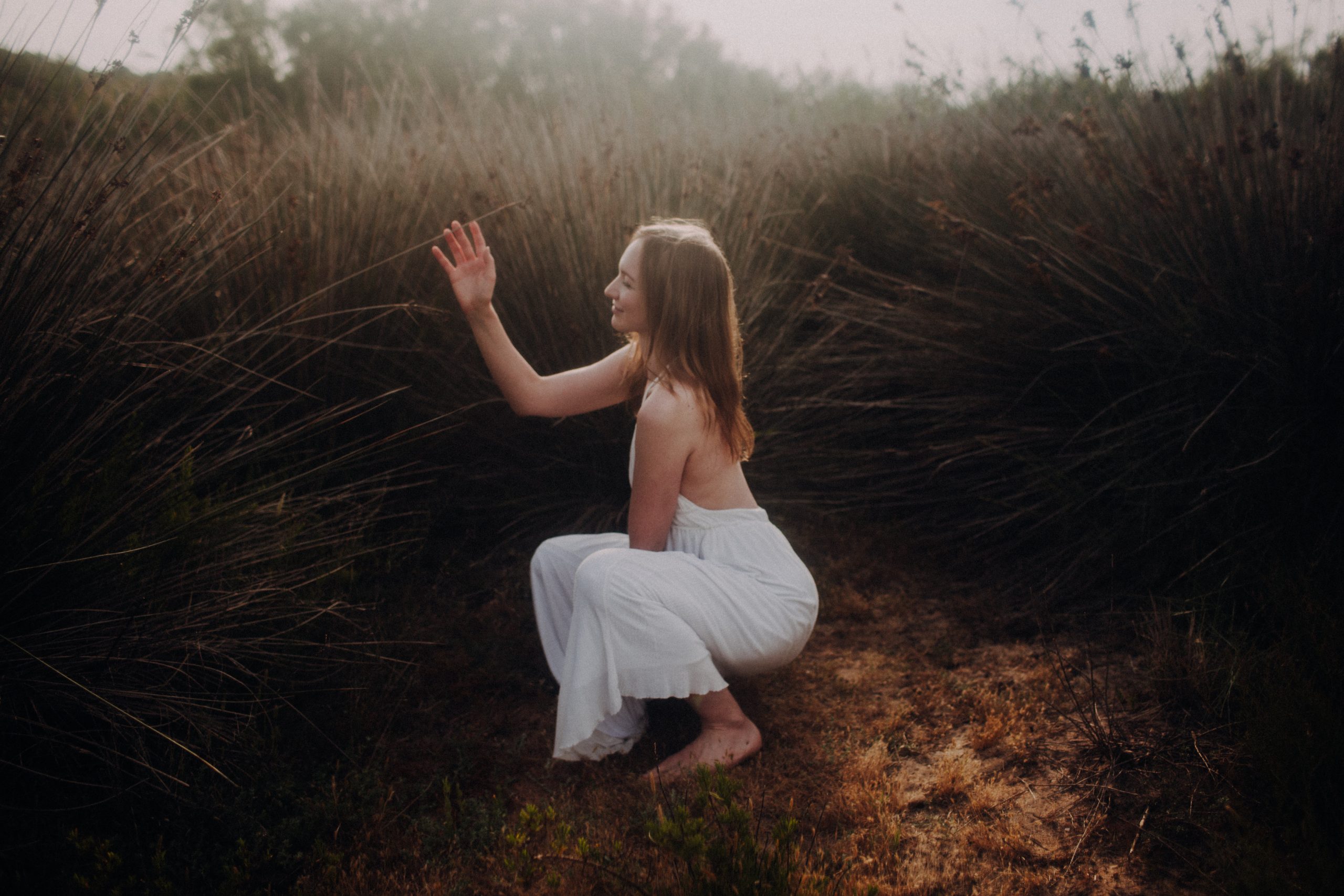 Boudoirfotoshooting zwischen Gräsern. Frau in weißem Kleid vor untergehender Sonne.