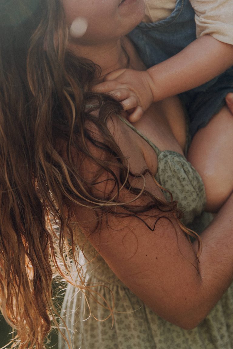 Details wie ein Kind sich an der Schulter der Mama festhält. Sie trägt ein sommerliches Kleid und hat Wassertropfen auf dem Arm.