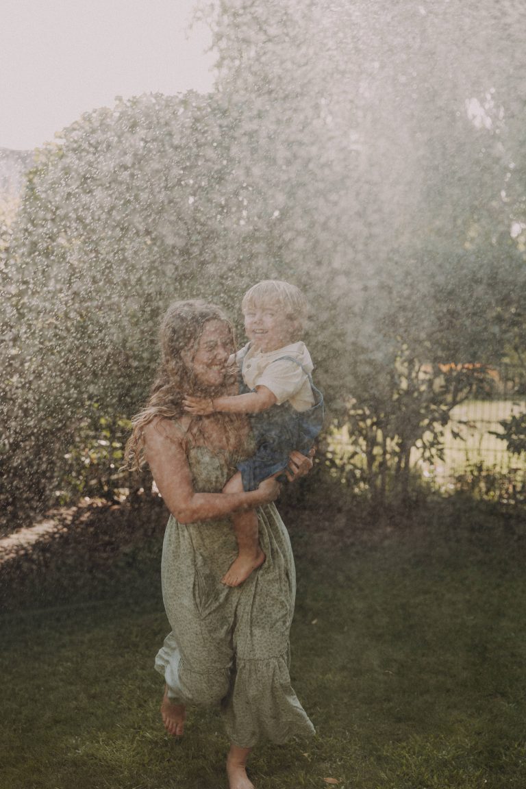 Mama mit Sohn auf dem Arm rennt durch die Tropfen eines Rasensprengers.
