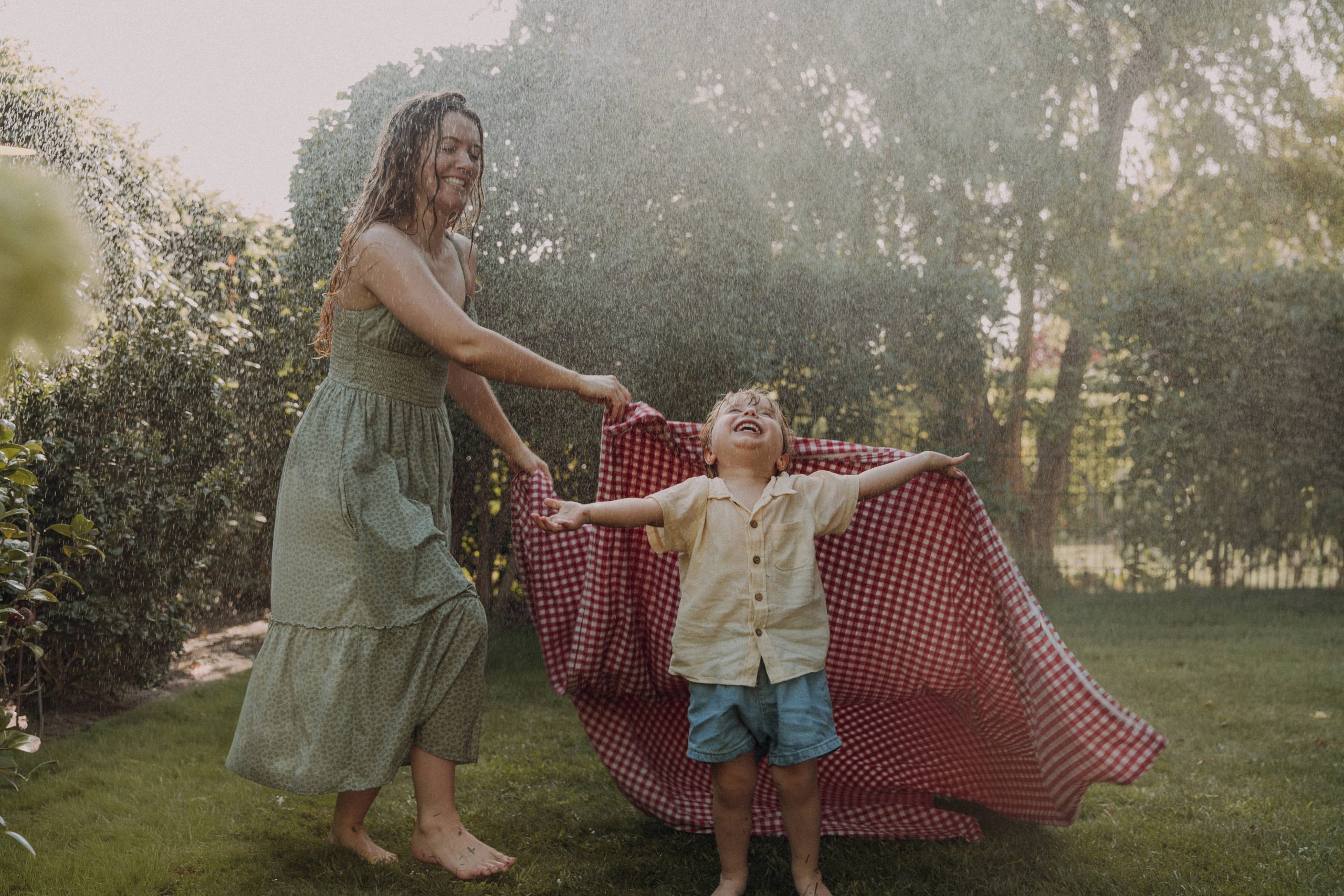 Fotos von einer Mama mit ihrem Kind im Garten unter einem Rasensprenger mit Sonne im Hintergrund