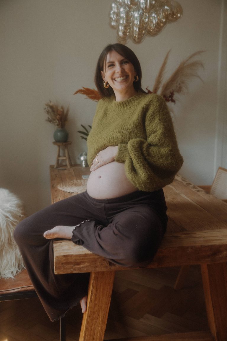 Schwangere Frau in einer gemütlichen Wohnung, sitzt auf einem Echtholztisch mit grünem Crop-Pullover.