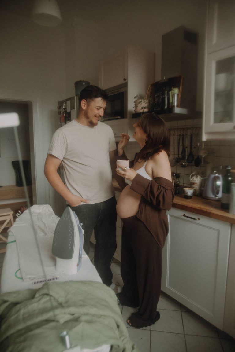Schwangeres Paar steht in Altbauküche und lächelt sich an. Man sieht ihren Babybauch und ein Bügeleisen, das als cooles Stilmittel wirkt.