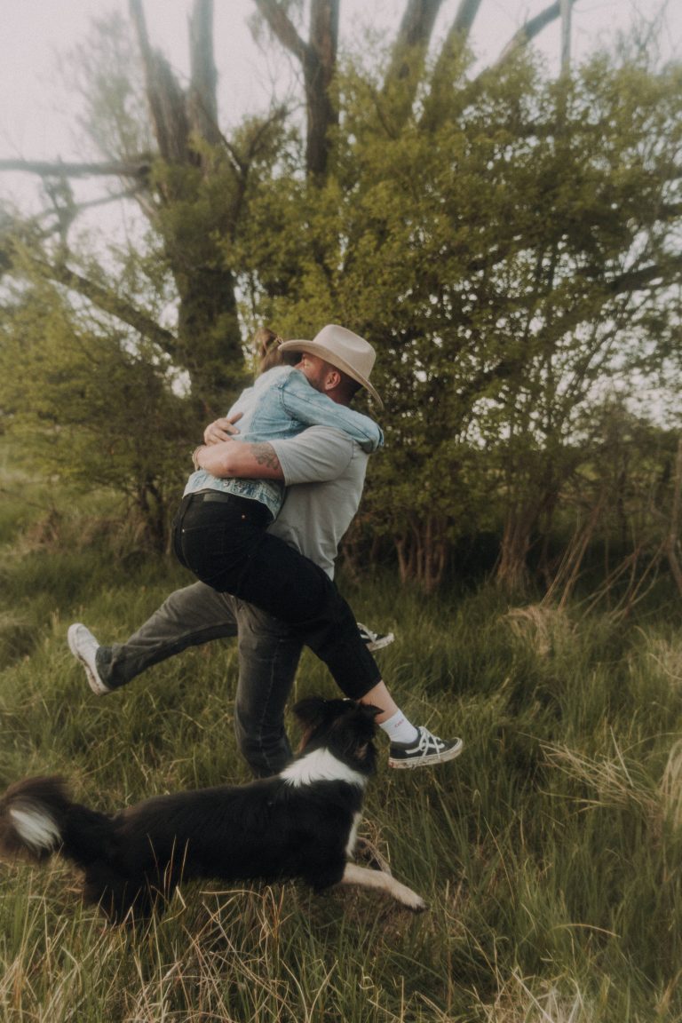 Ein Pärchen in der Natur. Er hat sie im Arm und dreht sie herum, der Hund der beiden rennt um sie herum.