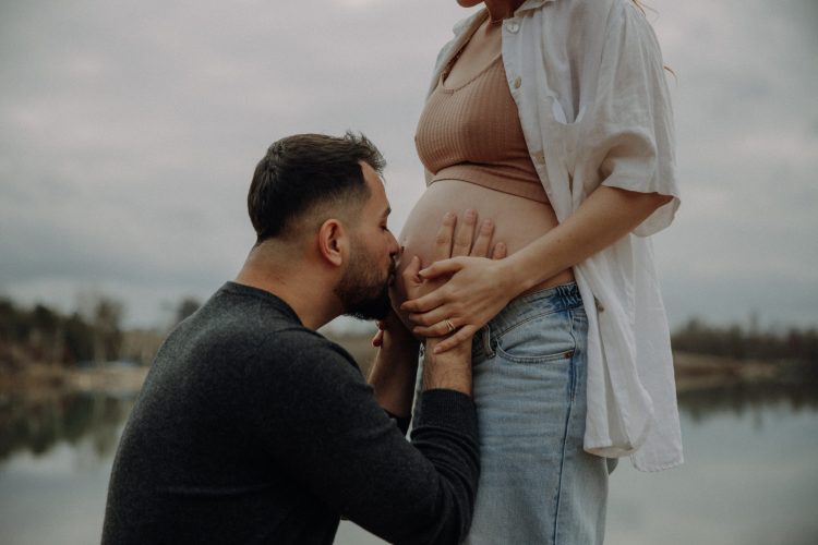 Mann küsst seiner schwangeren Frau auf den Bauch. Liebevolles Schwangerschaftsshooting in der Natur.