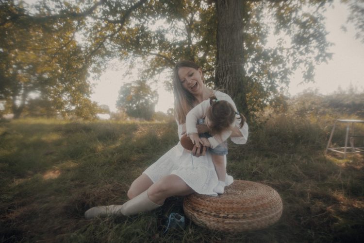 Authentisches Mutter-Kind Shooting Outdoor – Mutter lacht mit Tochter auf Rattanhocker unter spätsommerlichem Baum