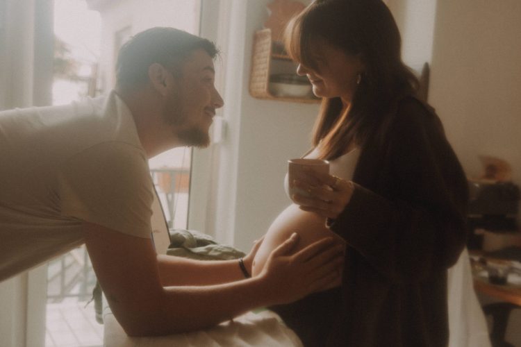 Ein schwangeres Paar bei einer Fotohomestory in der eigenen Küche. Er lehnt sich über ein Bügeleisen, hält ihren Bauch liebevoll in der Hand und lächelt ihr zu. Sie hat eine Kaffeetasse in der Hand.