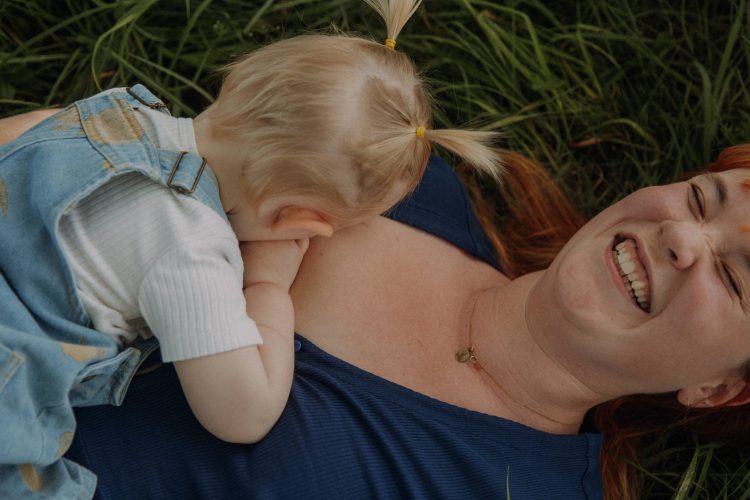 Mama-Kind Fotoshooting im Freien. Mama liegt lachend auf dem Rücken im Gras, Kind trinkt an ihrer Brust.